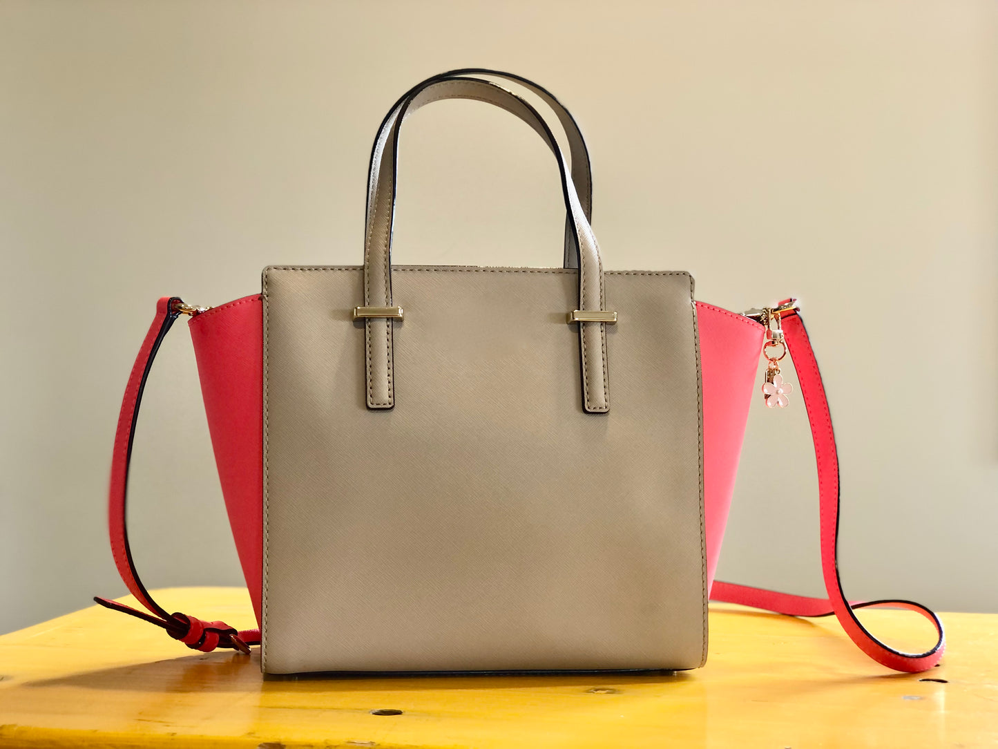 A beige and pink handbag sitting on a table. The handbag has a pink enamel blossom charm attached to one of the handles.