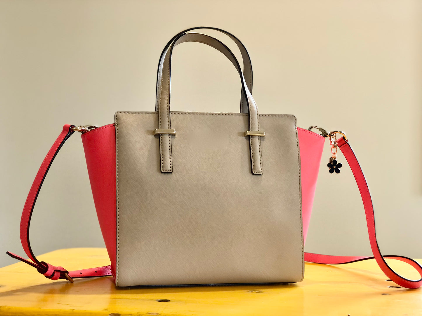 A beige and pink handbag sitting on a table. The handbag has a black enamel blossom charm attached to one of the handles.