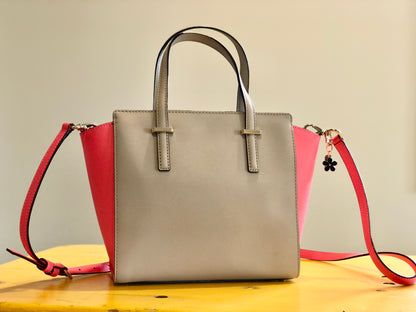 A beige and pink handbag sitting on a table. The handbag has a black enamel blossom charm attached to one of the handles.