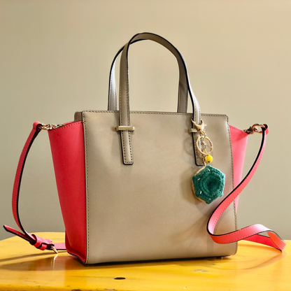 A beige and pink handbag sitting on a table. The handbag has a crocheted green donut charm attached to one of the handles.