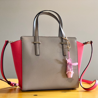 A beige and pink handbag sitting on a table. The handbag has a pink ballet slipper charm attached to one of the handles.