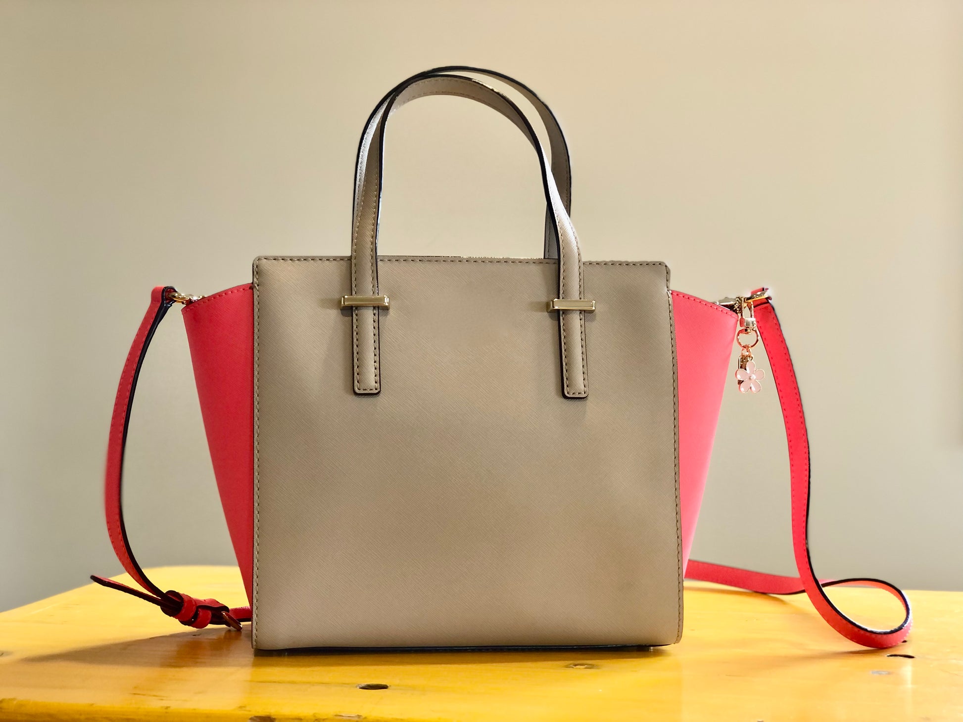 A beige and pink handbag sitting on a table. The handbag has a pink enamel blossom charm attached to one of the handles.