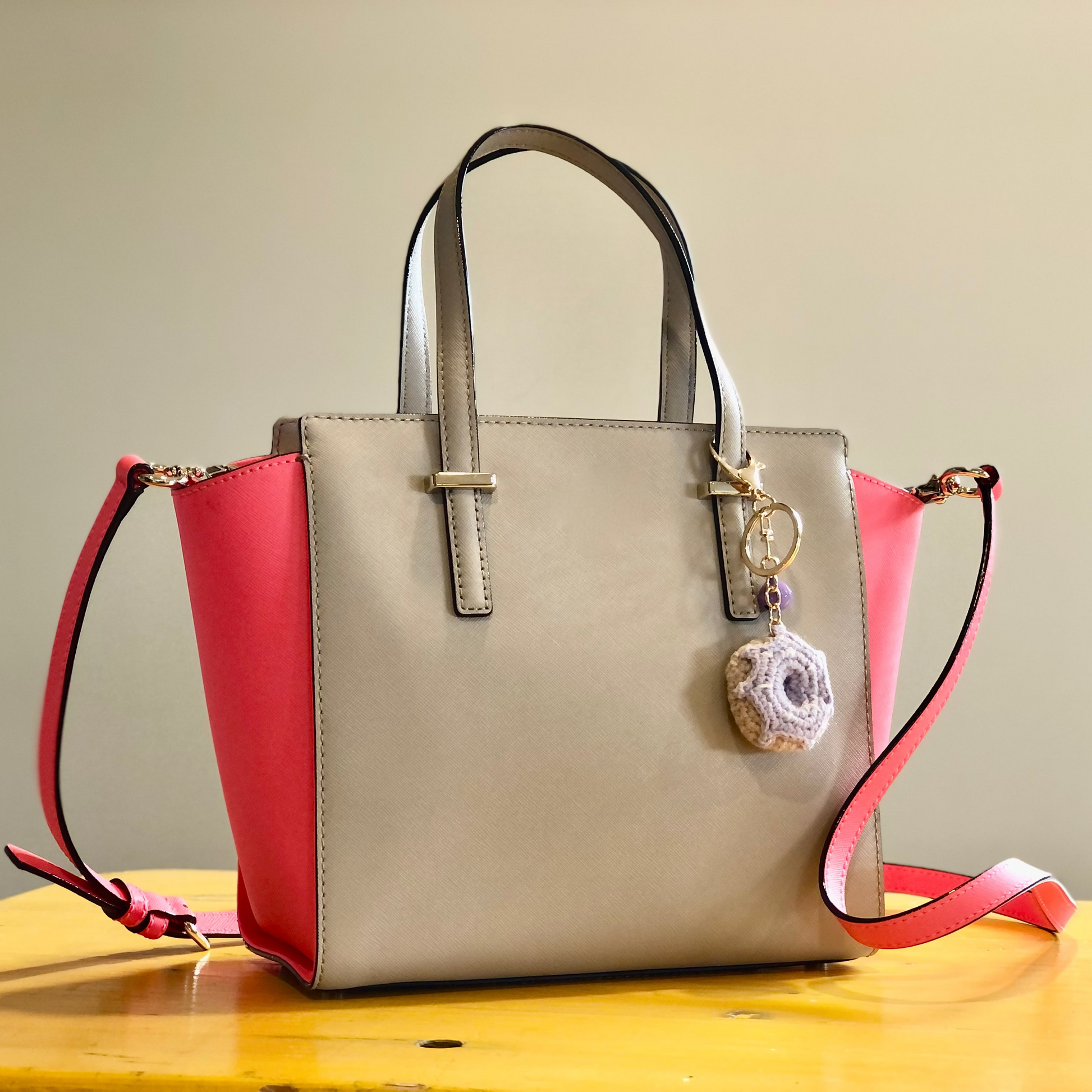 A beige and pink handbag sitting on a table. The handbag has a crocheted purple donut charm attached to one of the handles.