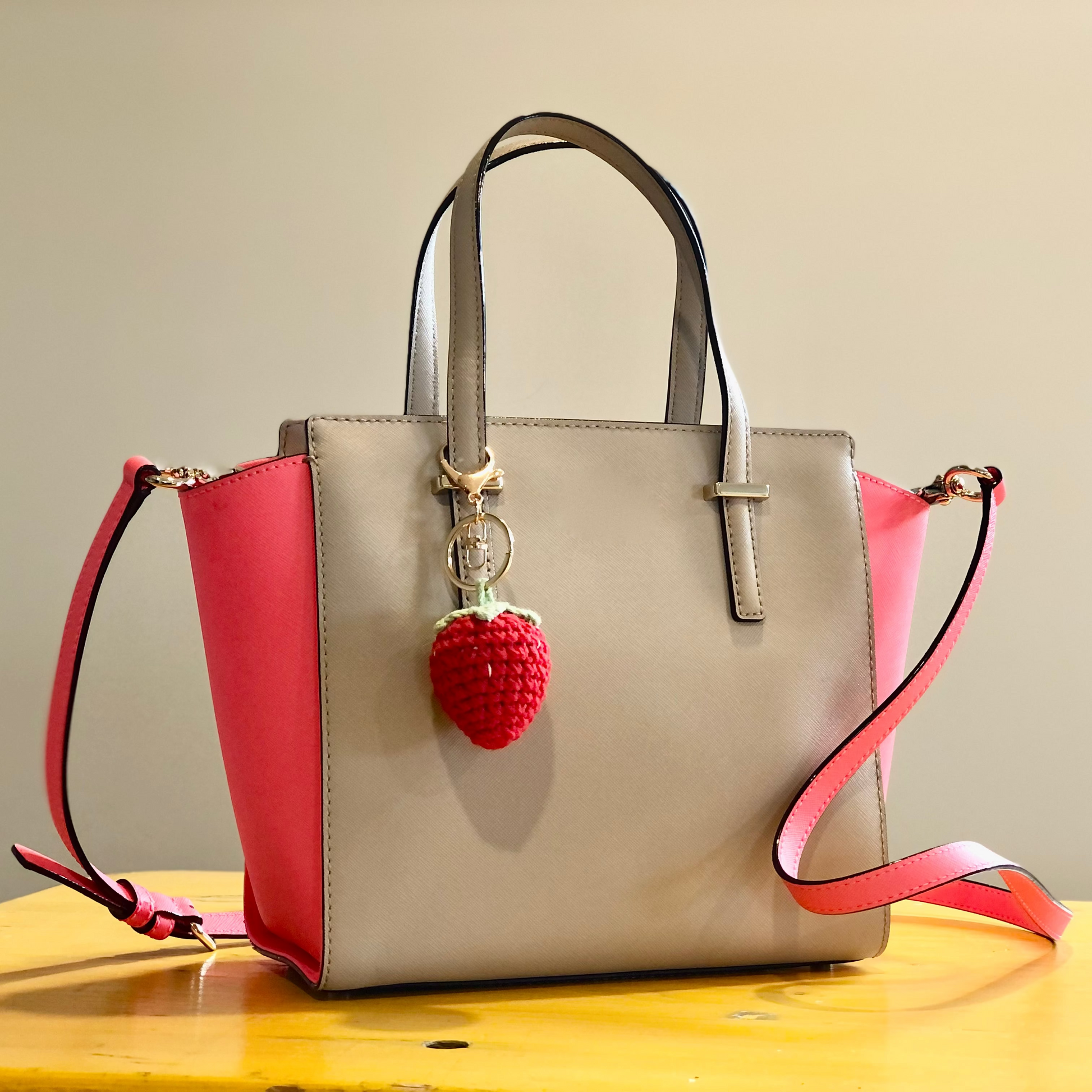 A crocheted handbag charm in the shape of a strawberry attached to a handbag.