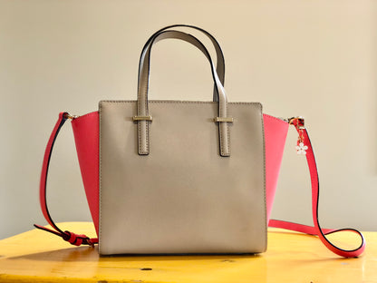 A beige and pink handbag sitting on a table. The handbag has a white enamel blossom charm attached to one of the handles.
