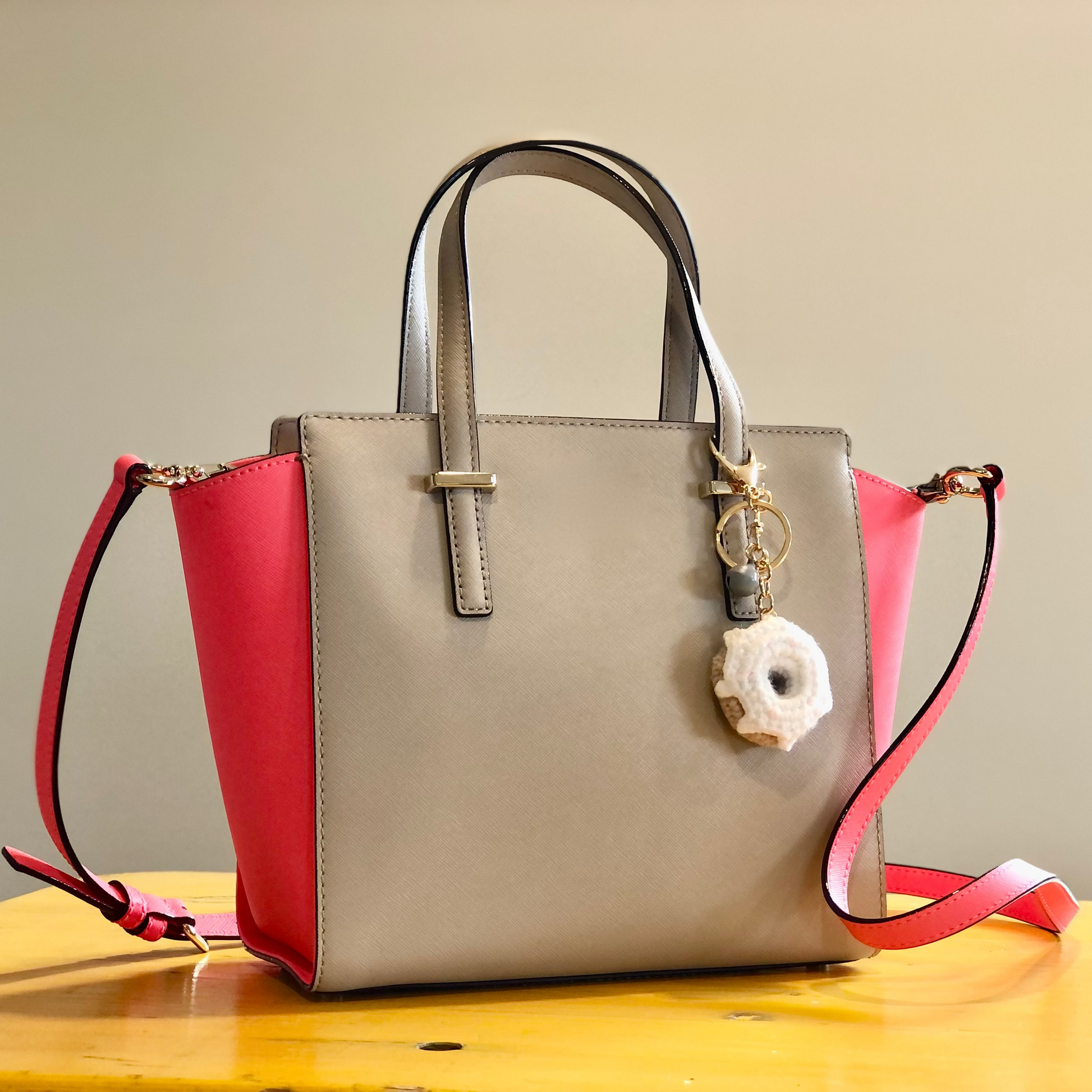 A beige and pink handbag sitting on a table. The handbag has a crocheted white donut charm attached to one of the handles.