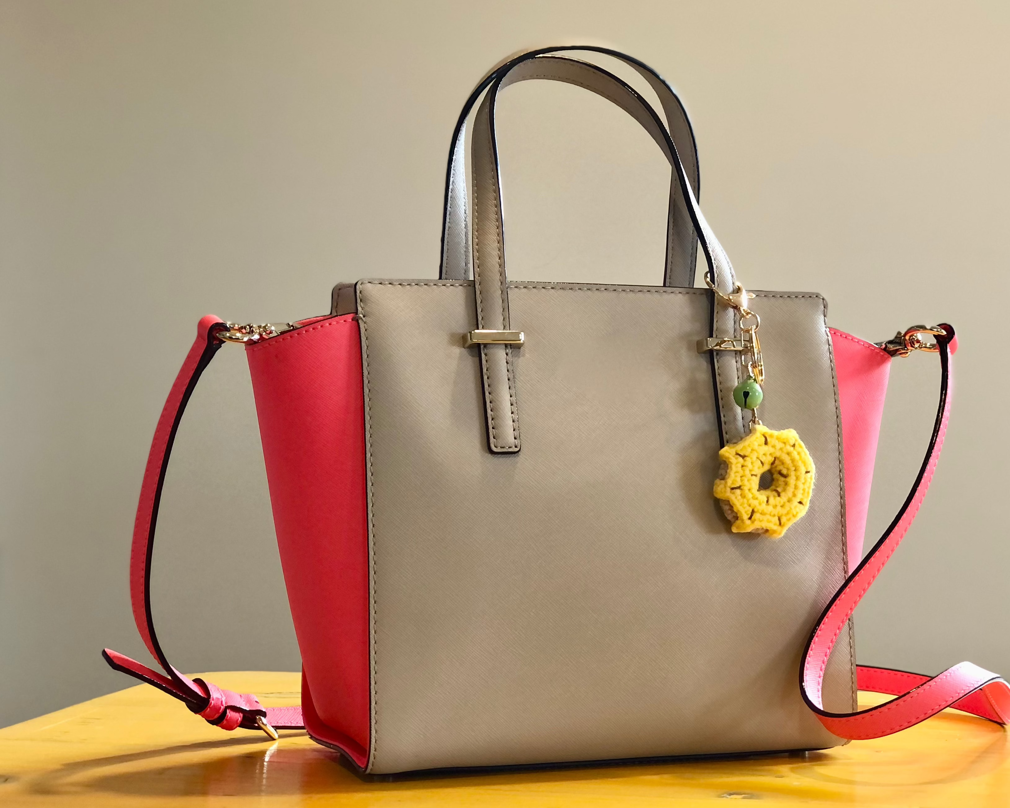 A beige and pink handbag sitting on a table. The handbag has a crocheted yellow donut charm attached to one of the handles.