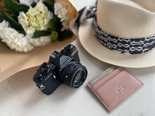 The Blossom (pink) Dark Hampton Leather Cardholder is displayed on a table with a hat, flowers, and camera.